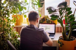 Man Works With Laptop Remotely From Home. Distant Work Place With Green Nature Inspired Home Office